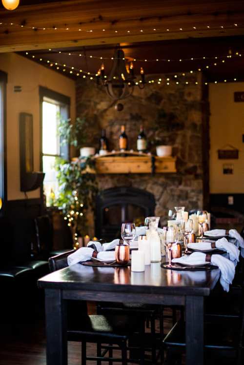 A cozy dining room with a long table set for a meal, candles, and warm lighting, featuring a stone fireplace in the background.