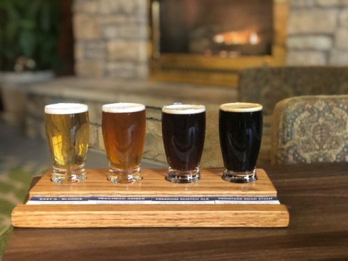 A wooden flight board holds four glasses of different beers in front of a cozy fireplace.