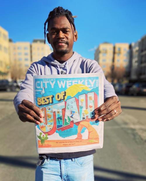 A person holds up a newspaper titled "City Weekly: Best of Utah" in a sunny urban setting.