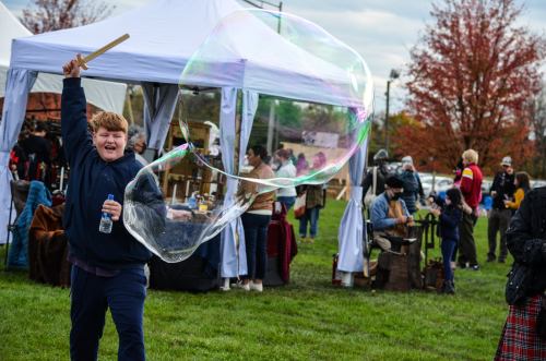 A boy joyfully swings a stick, creating a large bubble at an outdoor event with tents and people in the background.