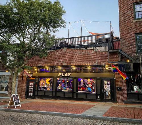 A vibrant bar with colorful window displays, a rainbow flag, and string lights on the rooftop in a brick building.