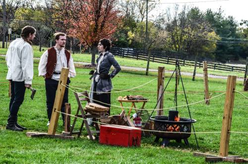 Three people in historical attire gather around a campfire in a grassy field, with trees and a fence in the background.