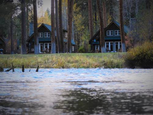 Two cozy cabins by a river, surrounded by tall trees and autumn foliage. Calm water reflects the serene landscape.
