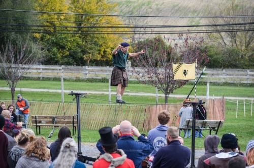 A performer in a kilt balances on a tightrope while an audience watches in a scenic outdoor setting.