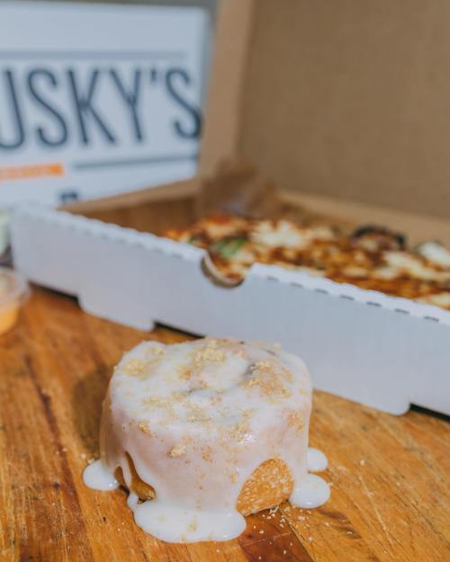 A cinnamon roll with icing sits on a wooden surface, with a pizza box in the background.