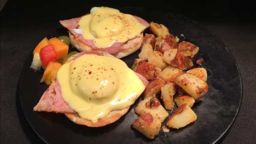 A black plate with two eggs Benedict on English muffins, ham, hollandaise sauce, and a side of crispy potatoes and fruit.