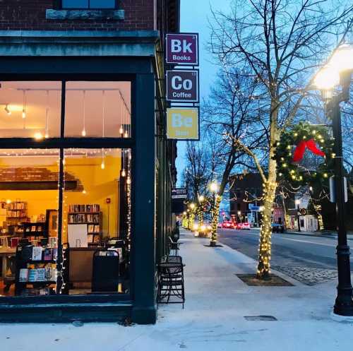 A cozy bookstore and coffee shop at dusk, with festive decorations and warm lights illuminating the street.