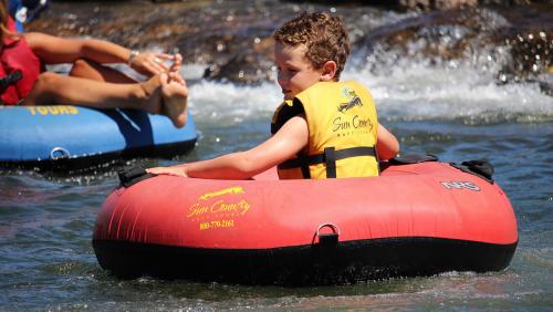 A young boy in a red inflatable tube enjoys floating on a river, wearing a yellow life jacket.