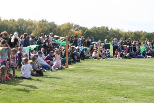 A large crowd of people sitting on grass, enjoying an outdoor event with colorful decorations in the background.