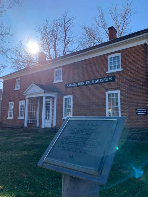 A brick building with a sign reading "Amana Heritage Museum," featuring a plaque in the foreground and sunlight shining.