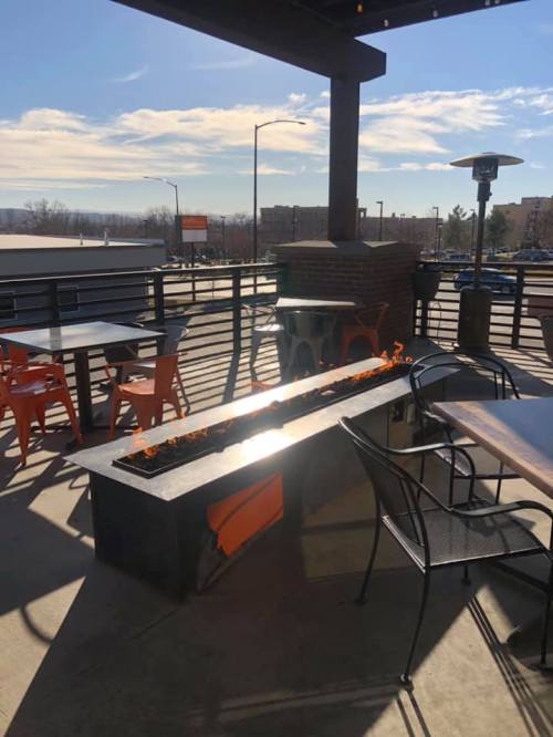 A rooftop patio with a fire pit, tables, and chairs, under a clear blue sky with scattered clouds.
