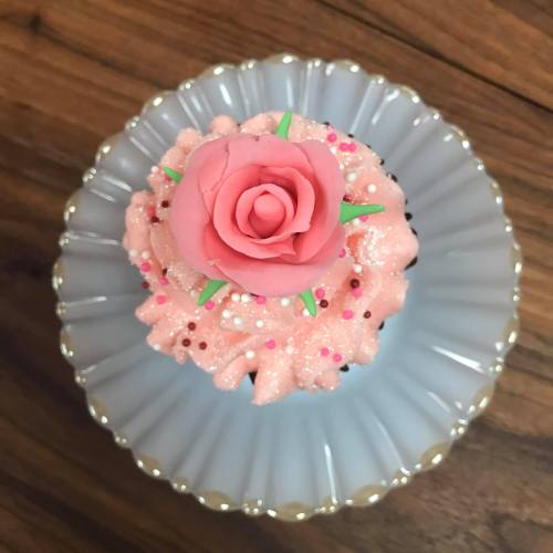 A pink frosted cupcake topped with a rose decoration and colorful sprinkles, served on a decorative plate.
