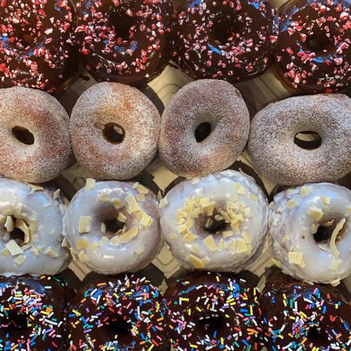 A variety of colorful donuts arranged in rows, featuring sprinkles, chocolate glaze, and powdered sugar.