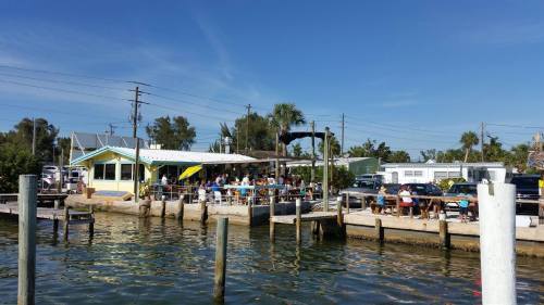 A waterfront restaurant with outdoor seating, surrounded by palm trees and a clear blue sky. Boats are docked nearby.
