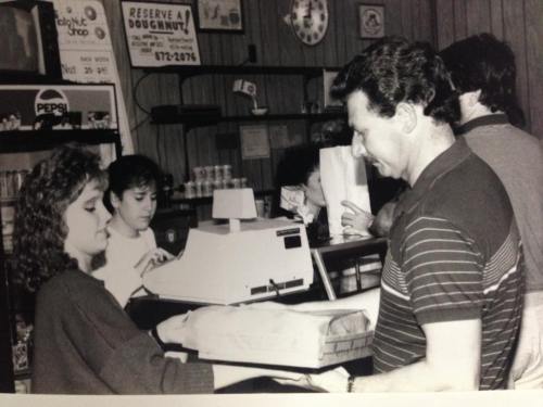 A black-and-white photo of a busy shop with a cashier serving a customer, while others wait in line.