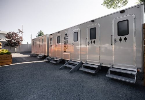 Row of portable restroom trailers parked on gravel, featuring signs for men's and women's facilities.