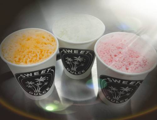 Three cups of colorful shaved ice in orange, white, and pink, labeled "Sweet Dulce," on a shiny surface.