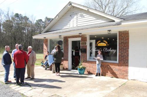 A group of people socializing outside a bakery with a sign, surrounded by trees and a clear sky.