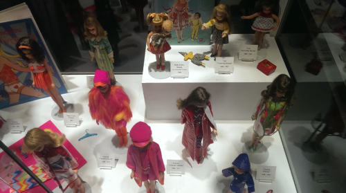 A display of vintage dolls in various outfits, showcasing different styles and accessories on a white pedestal.