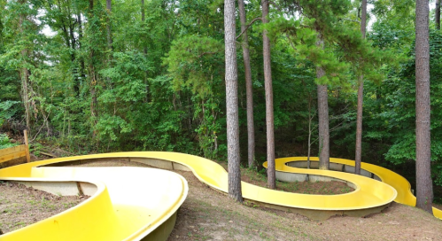 A winding yellow waterslide surrounded by tall trees in a wooded area.