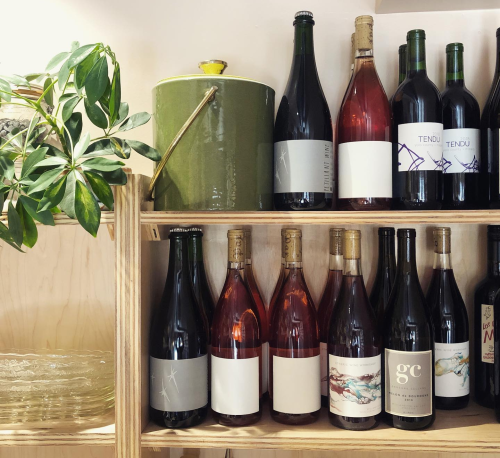 A wooden shelf displaying various wine bottles, including reds and rosés, alongside a green container and a plant.