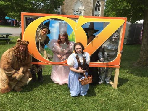 A group of people dressed as characters from "The Wizard of Oz" pose in front of a large "OZ" sign outdoors.