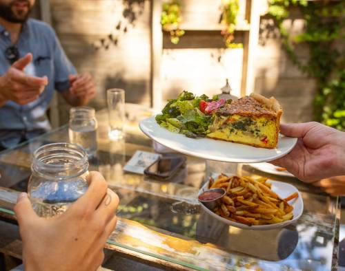 A hand serves a slice of quiche with salad, alongside a plate of fries, at a sunny outdoor table.