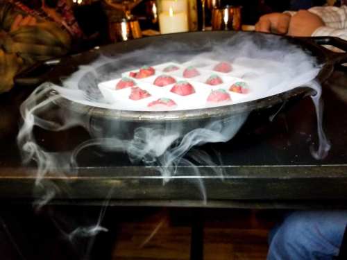 A black pot with strawberries on a white tray, surrounded by swirling smoke, set on a table.