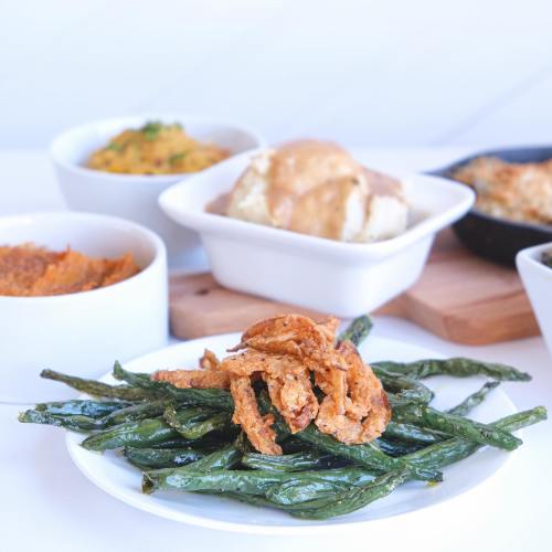 A plate of crispy green beans topped with fried onions, surrounded by bowls of various side dishes.