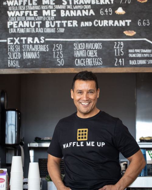 A smiling man in a black "Waffle Me Up" shirt stands in front of a menu board at a waffle shop.