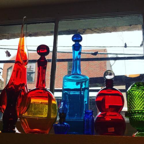 Colorful glass bottles in various shapes, displayed on a windowsill with a view of a brick building outside.