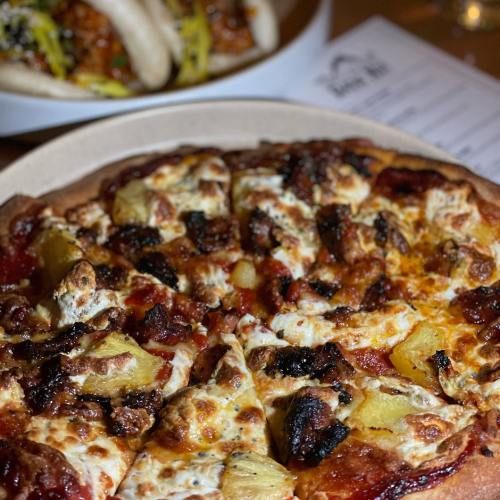 A close-up of a pizza topped with cheese, pineapple, and charred edges, with another dish blurred in the background.