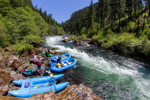 A group of people preparing kayaks and rafts by a river surrounded by lush green trees and rocky terrain.