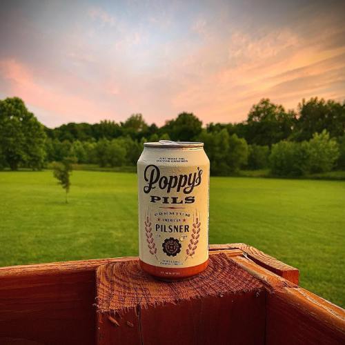 A can of Poppy's Pils beer sits on a wooden railing, with a lush green field and sunset sky in the background.