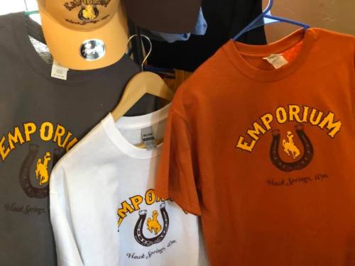 Three t-shirts in gray, white, and orange hanging on a rack, all featuring the word "EMPORIUM" and a horse logo.