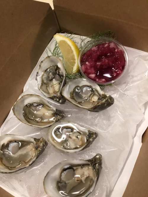 A takeout box containing fresh oysters on ice, a lemon wedge, and a small cup of red sauce.