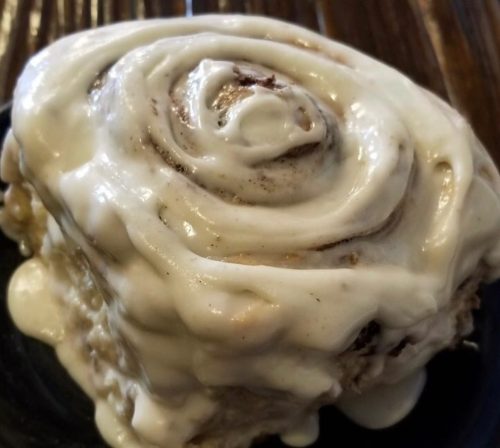 A close-up of a cinnamon roll topped with creamy white icing on a dark plate.