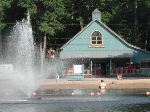 A blue building with a peaked roof near a lake, featuring a fountain and a sandy beach area.