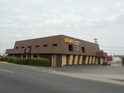 A brown building with a sloped roof, featuring a sign that reads "FAME" and surrounded by greenery and a parking lot.