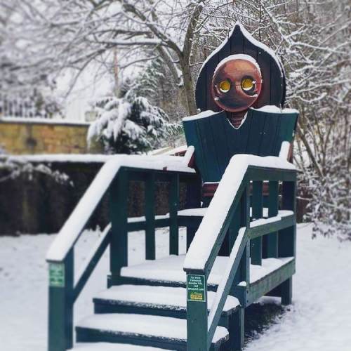 A large, whimsical wooden figure with big eyes sits on a snowy staircase in a winter landscape.