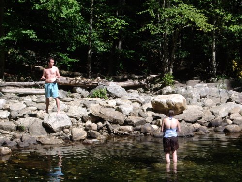 Two people by a rocky riverbank; one stands on a rock while the other wades in the water, surrounded by trees.