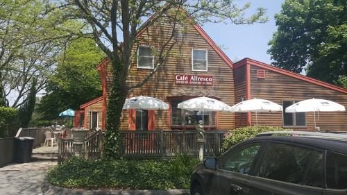 A cozy café with a wooden exterior, outdoor seating, and umbrellas, surrounded by trees on a sunny day.