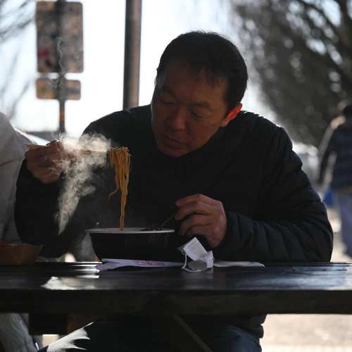 A man sits at a table, enjoying a steaming bowl of noodles on a sunny day.