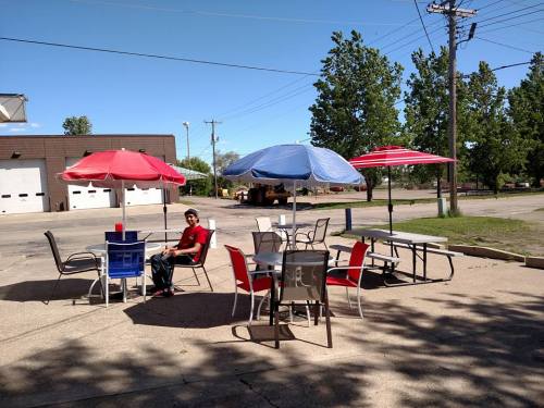 A man sits at a table under colorful umbrellas in an outdoor seating area with trees and a clear blue sky.