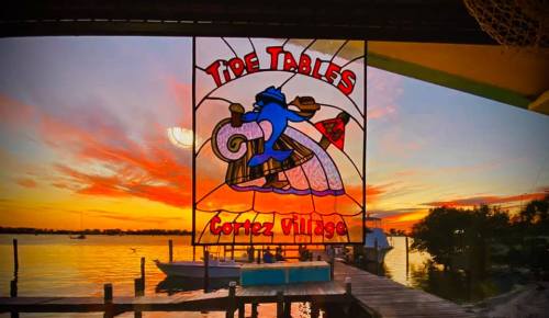 Colorful sunset over water with a stained glass sign reading "Tide Tables, Cortez Village" in the foreground.