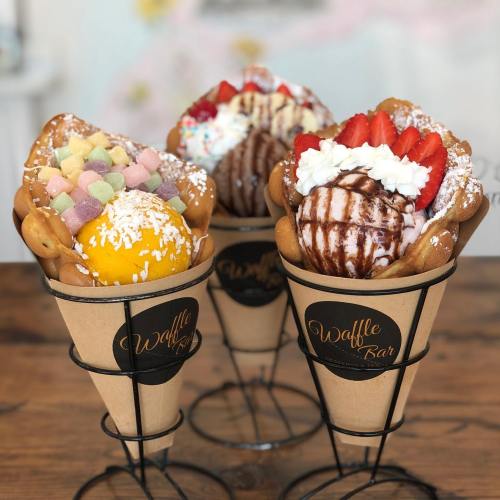 Three waffle cones filled with ice cream, toppings, and colorful sweets, displayed in a wire holder.
