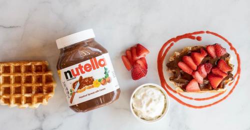 A jar of Nutella beside a waffle topped with strawberries and whipped cream, with a strawberry sauce drizzle on a marble surface.