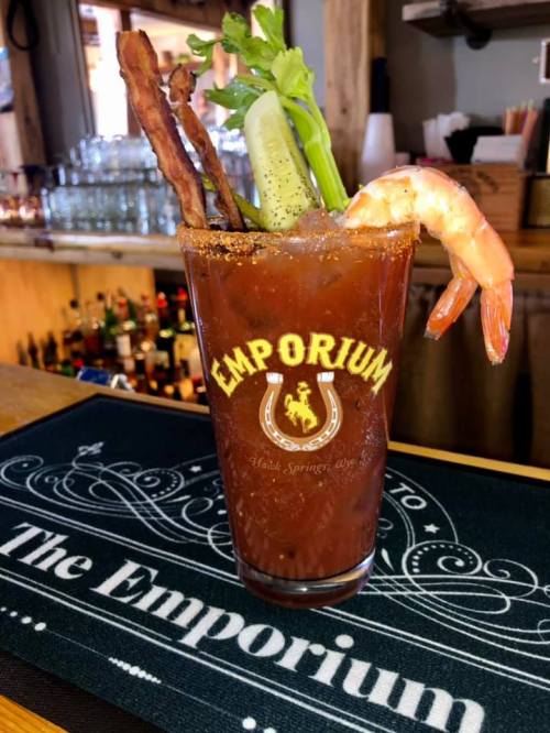 A cocktail glass filled with a Bloody Mary, garnished with celery, bacon, and a shrimp, on a bar mat.