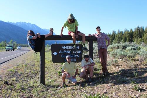 A group of five friends in colorful shirts poses playfully around an American Alpine Club sign in a scenic outdoor setting.