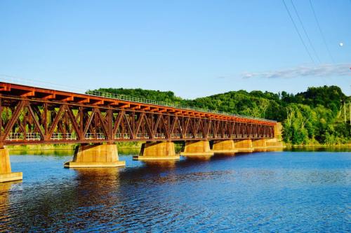 A rust-colored bridge spans a calm river, surrounded by lush green hills under a clear blue sky.
